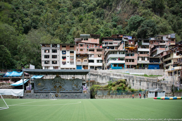 Green sports field below closely packed multistory hillside buildings and forested mountains in Aguas Calientes.