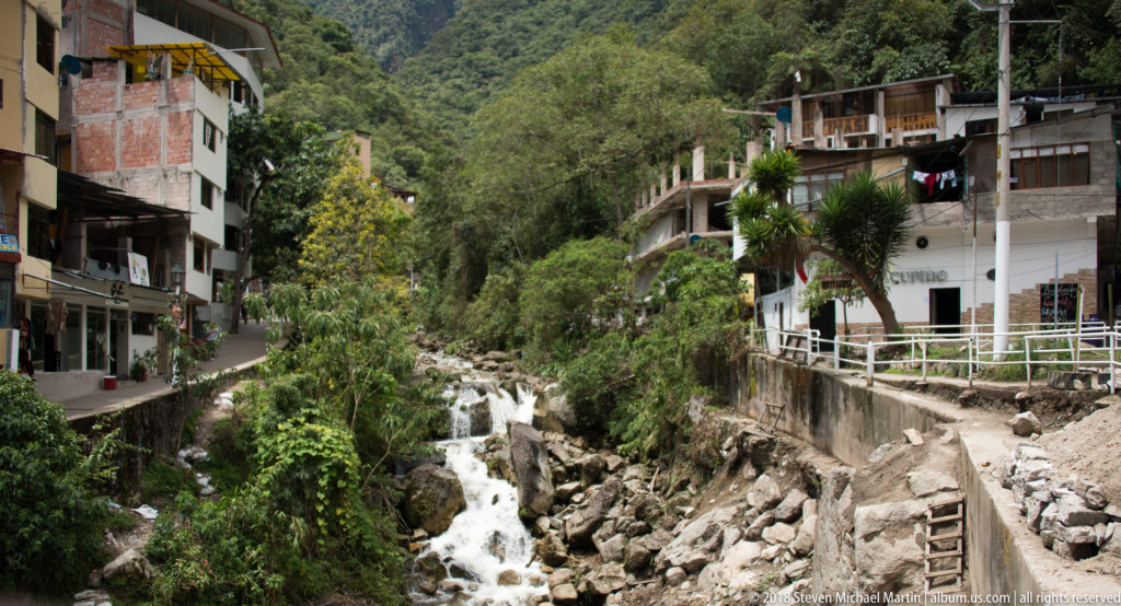 Rocky mountain stream cascading between buildings and retaining walls in the steep green valley of Aguas Calientes.