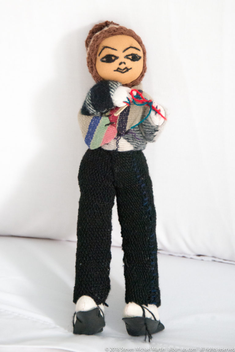 Handmade textile doll in black trousers and patterned Andean-style clothing standing against a white background.