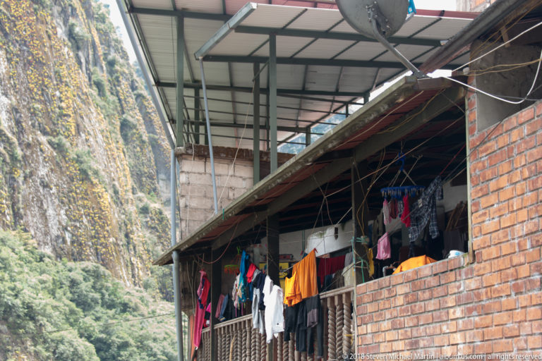Laundry hangs from a brick residential balcony beneath metal roofing, with a steep forested cliff rising immediately behind.