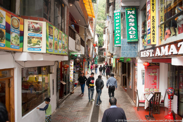 Narrow pedestrian street crowded with restaurant, hotel, and money-exchange signs as people walk between closely spaced buildings.