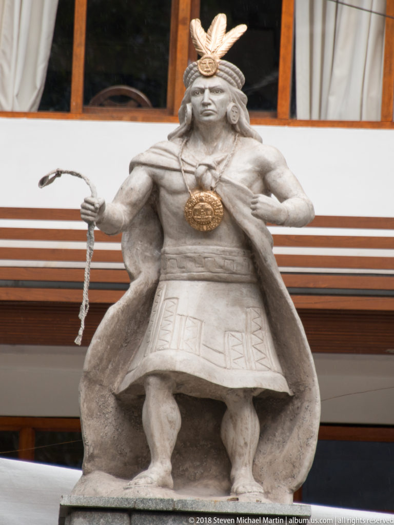Standing Inca-themed stone figure with feathered headdress, circular chest ornament, cape, and long staff or cord.