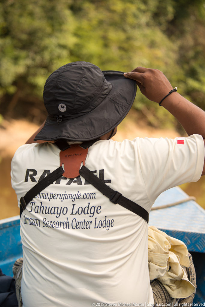 Canoe passenger seen from behind adjusting a black sun hat, wearing a shirt printed with Tahuayo Lodge and Amazon Research Center Lodge.