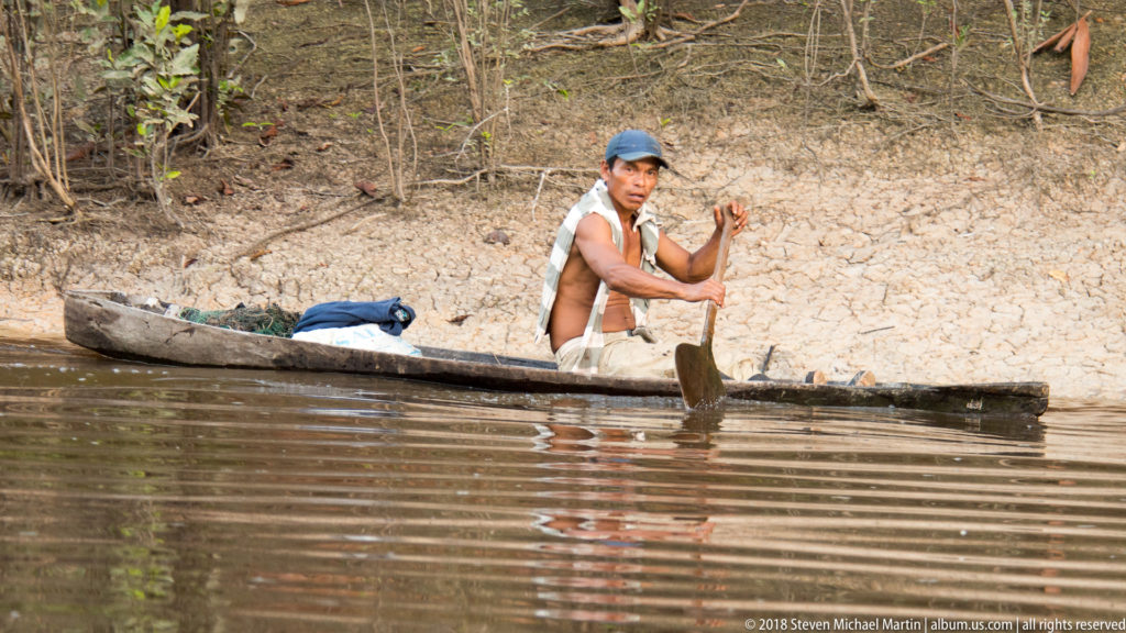 Man paddling a long narrow wooden dugout canoe beside a dry, cracked riverbank edged by forest.