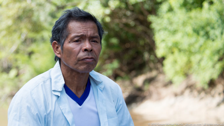 Close portrait of an older man in a pale shirt over a blue-and-white top, with blurred green foliage behind.