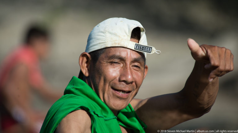 Smiling canoe paddler raising one hand in a close portrait, with green cloth over his shoulders and a blurred figure behind.