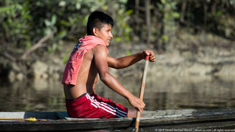 Close side view of a person paddling a narrow wooden canoe, with the paddle held vertically against a softly blurred riverbank.