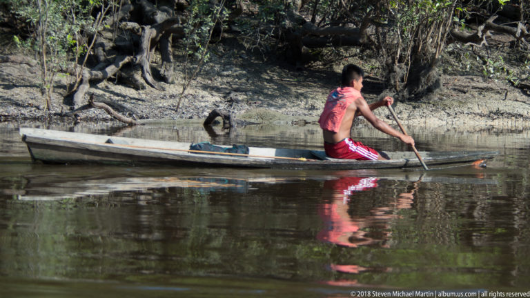 Person paddling a narrow wooden canoe through a dark forest-lined Amazonian channel beside exposed roots and muddy bank.