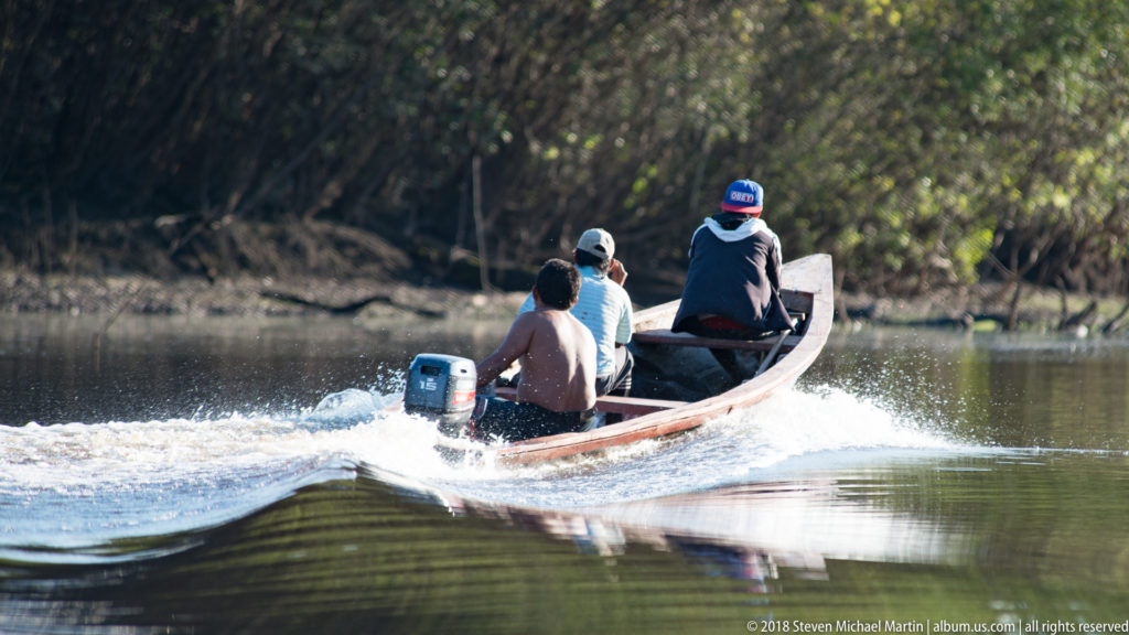 Three people travel away in a narrow wooden motor canoe, leaving a white wake across a calm forest-lined Amazon waterway.
