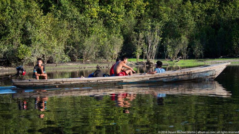 Several people seated in a long wooden canoe on calm reflective water beside dense green Amazonian forest.