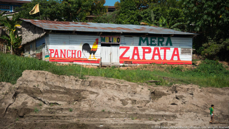 Wooden building painted MOLINO MERA, PANCHO, and ZAPATA with rooster emblem above a sandy eroded riverbank.