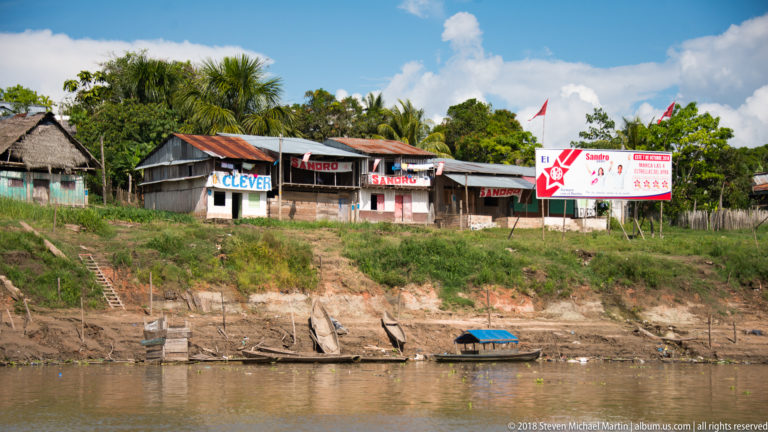 Riverside houses painted CLEVER and SANDRO stand above an eroded bank with campaign billboard and small boat below.