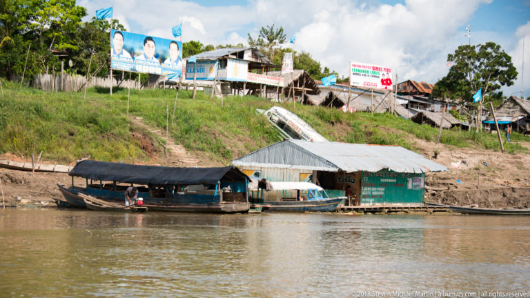 Covered riverboat and green waterfront building below a steep bank lined with houses and political signs.