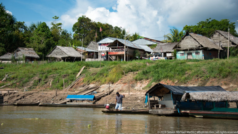 Timber and thatch-roofed houses stand above an eroded riverbank with small boats moored below.
