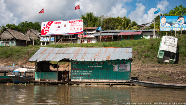 Green riverside shop marked MULTISERVICIOS CELIA below campaign signs on a steep bank with boats at the waterline.