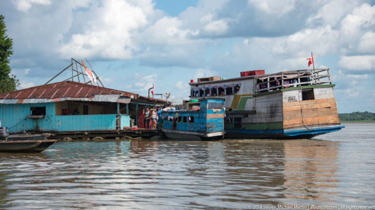 Two large multi-deck riverboats moored beside a low wooden waterfront building with people and flags nearby.