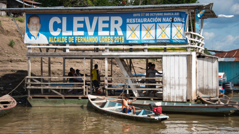 Stilted riverside building with large CLEVER mayoral campaign sign, people below, and a small motor canoe in front.