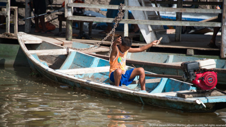 Man sits in a narrow wooden canoe beside an exposed red gasoline engine near a timber river landing.
