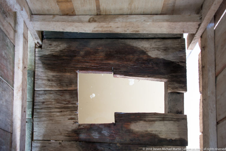 Irregular rectangular opening in weathered wooden planks reveals pale river water beyond a shadowed interior.