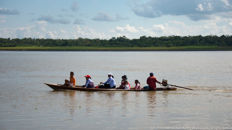 Long motorized canoe with several passengers crosses wide river water toward a low forested bank.
