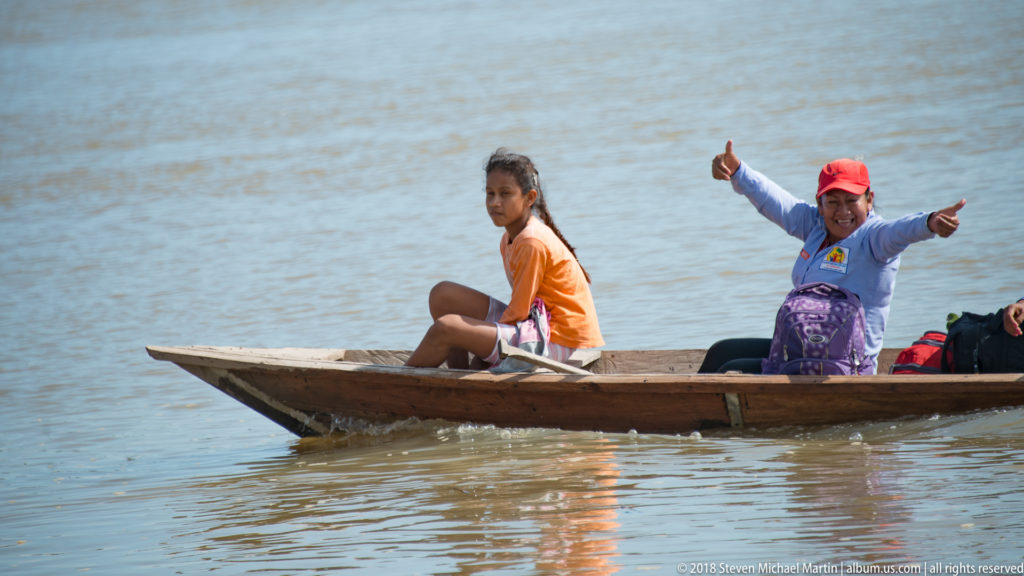 Woman in red cap raises both arms from a narrow wooden canoe while a younger passenger sits ahead.