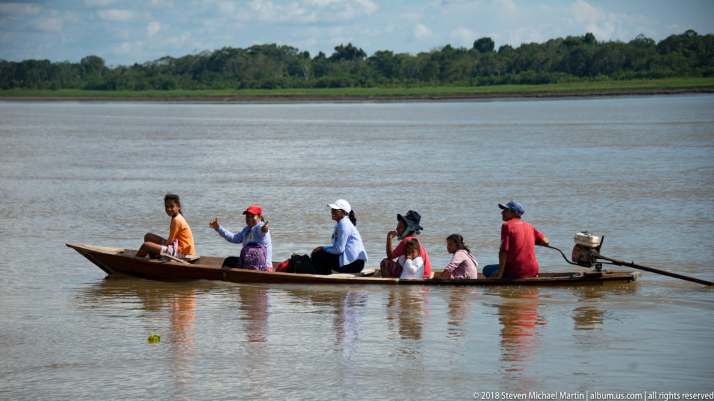 Several passengers sit in a long narrow motorized wooden canoe crossing broad brown river water.