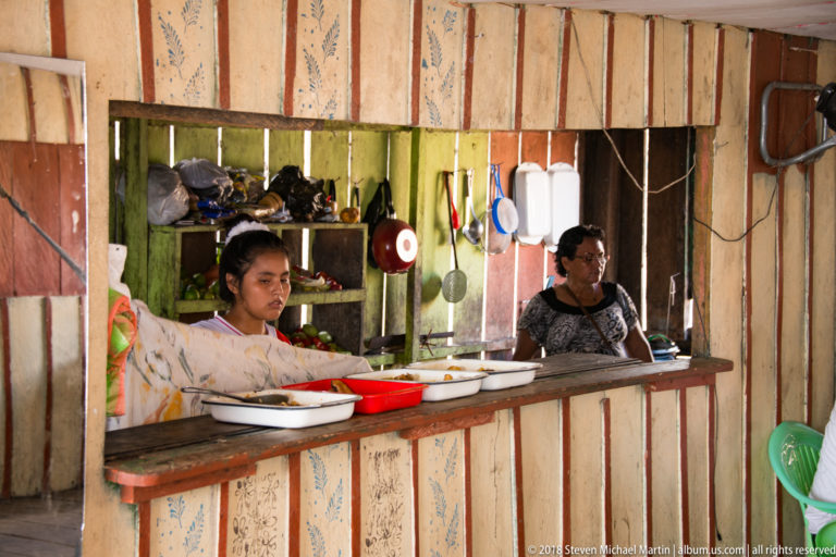 Two women behind a wooden food-stall counter with bowls, pans, utensils, shelves, and patterned wall boards.