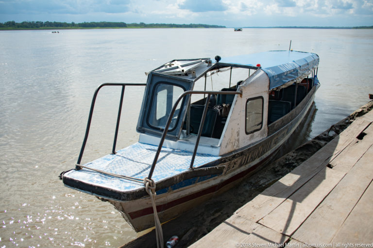 Compact cabin motorboat tied to a wooden dock with broad river water and forested shoreline beyond.