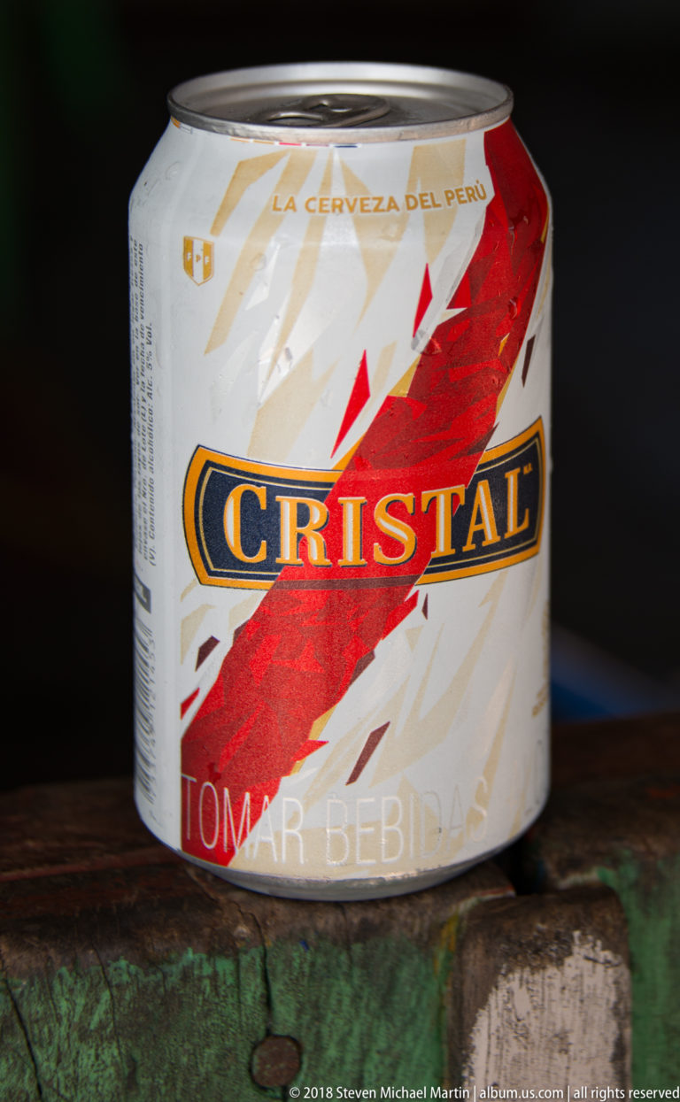 Squat Cristal beer can with red diagonal label and “La Cerveza del Perú” text resting on worn green-painted wood.