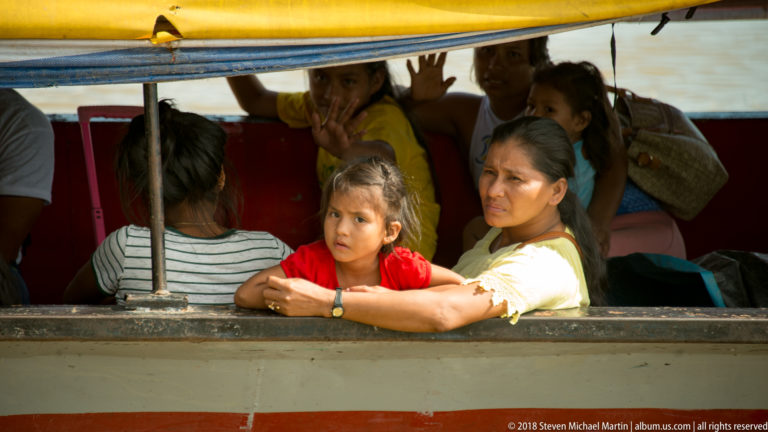 Woman holding a young child at an open riverboat window, with adults and children seated closely behind them.