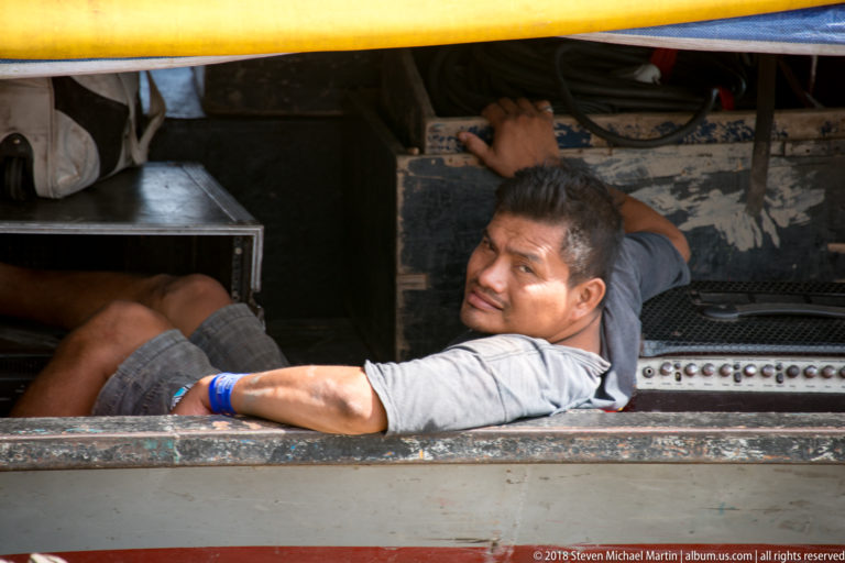 Man resting his arm on the metal side of a riverboat beside worn controls, cables, and equipment.