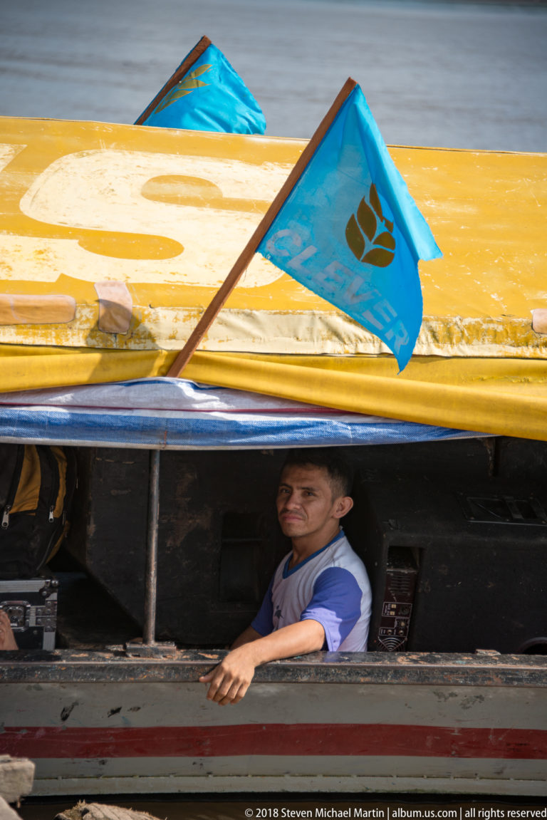 Man leaning from the open side of a yellow-canopied riverboat beneath blue flags.