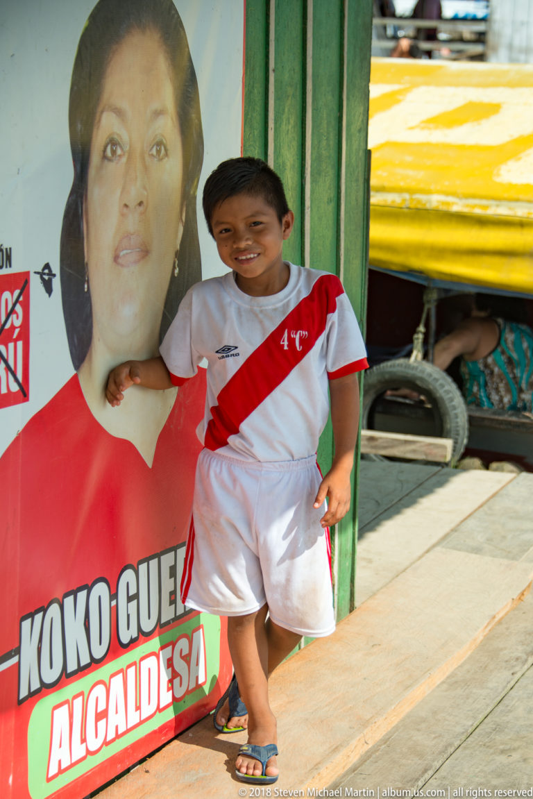 Boy in a white Peru football kit standing beside a large campaign poster reading “Koko Guerra Alcaldesa.”