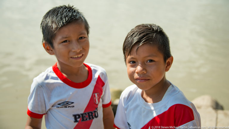 Two boys standing beside the river in white Peru football jerseys with red diagonal sashes.