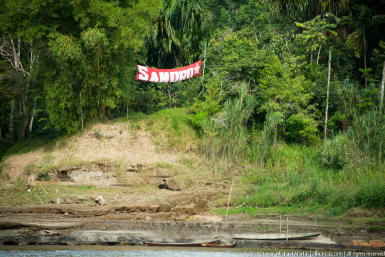 Red SANDRO banner hangs before dense tropical forest above a muddy riverbank and narrow boat.