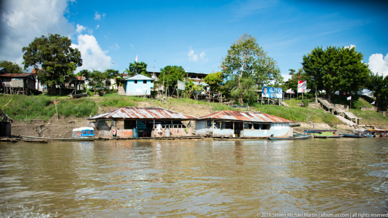 Low corrugated-roof floating structures and small boats at the Tamshiyacu shoreline, with houses and trees on the bank above.