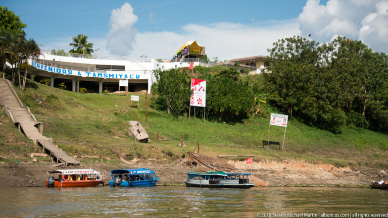 Welcome sign for Tamshiyacu above a steep grassy riverbank with wooden stairs and several narrow boats below.