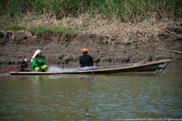 Two people ride in a long narrow wooden canoe beside a steep exposed muddy riverbank.