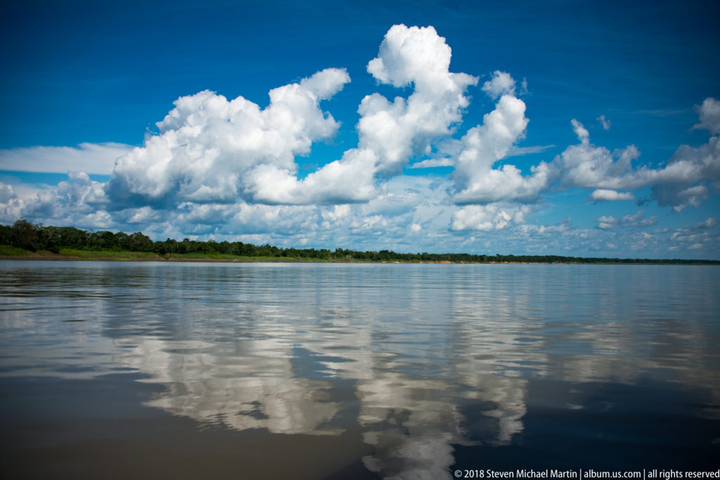 Broad calm Amazon River reflecting large white cumulus clouds, with a low forested horizon in the distance.