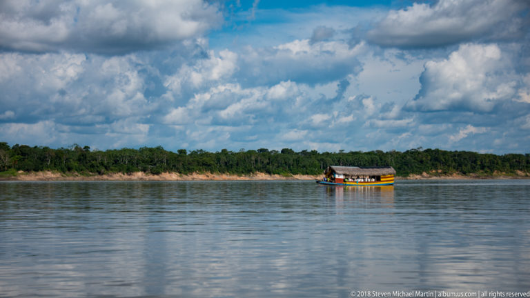 Small thatch-roofed passenger boat crossing broad Amazon River water near a distant forested bank beneath large clouds.