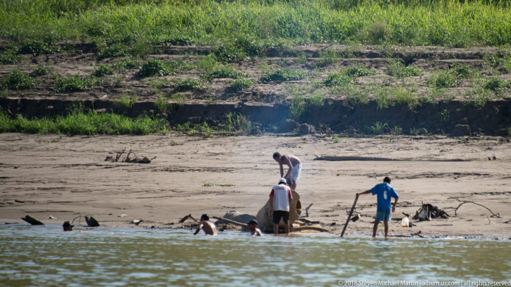 Several people stand and bend beside a low craft or equipment in shallow water along a sandy Amazon River bank.