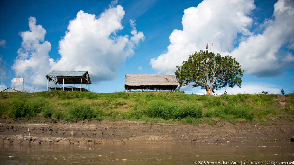 Open-sided riverside shelters and a broad tree on a grassy bank above muddy Amazon River water under towering clouds.