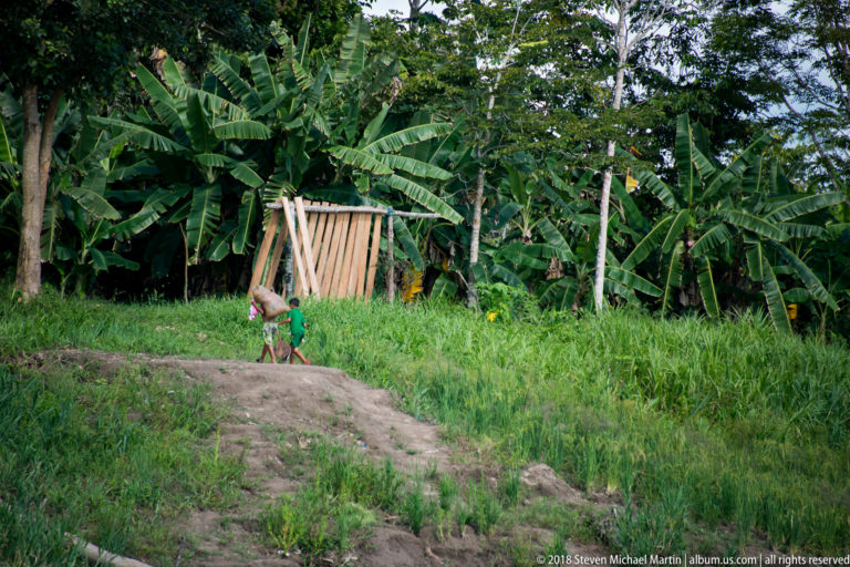 Small child walks beside dense banana plants and a simple upright wooden panel on a grassy riverbank rise.