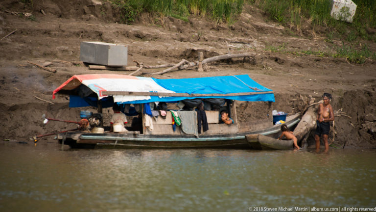 People beside a small wooden river craft covered with blue and white tarpaulins and hung with clothing and supplies.