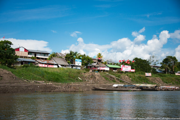 Wide riverside settlement with houses, campaign signs, grassy bank, and moored boats beneath a bright blue sky.