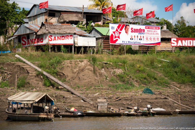 Wooden riverside buildings covered in red SANDRO signs and flags above narrow boats on the muddy shore.