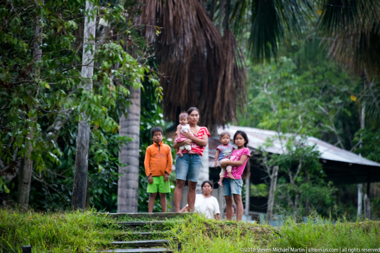 Adults and children standing together beside dense tropical vegetation and a simple roofed structure.