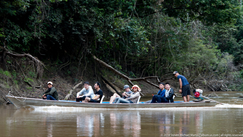 Group of passengers riding in a long narrow motorboat beside a densely forested riverbank.