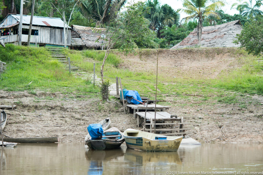 Small motorboats tied beside a muddy riverbank with wooden steps, simple buildings, and palms above.