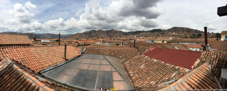 Wide rooftop view over Cusco’s red tiles toward Andean ridges beneath towering dramatic clouds.